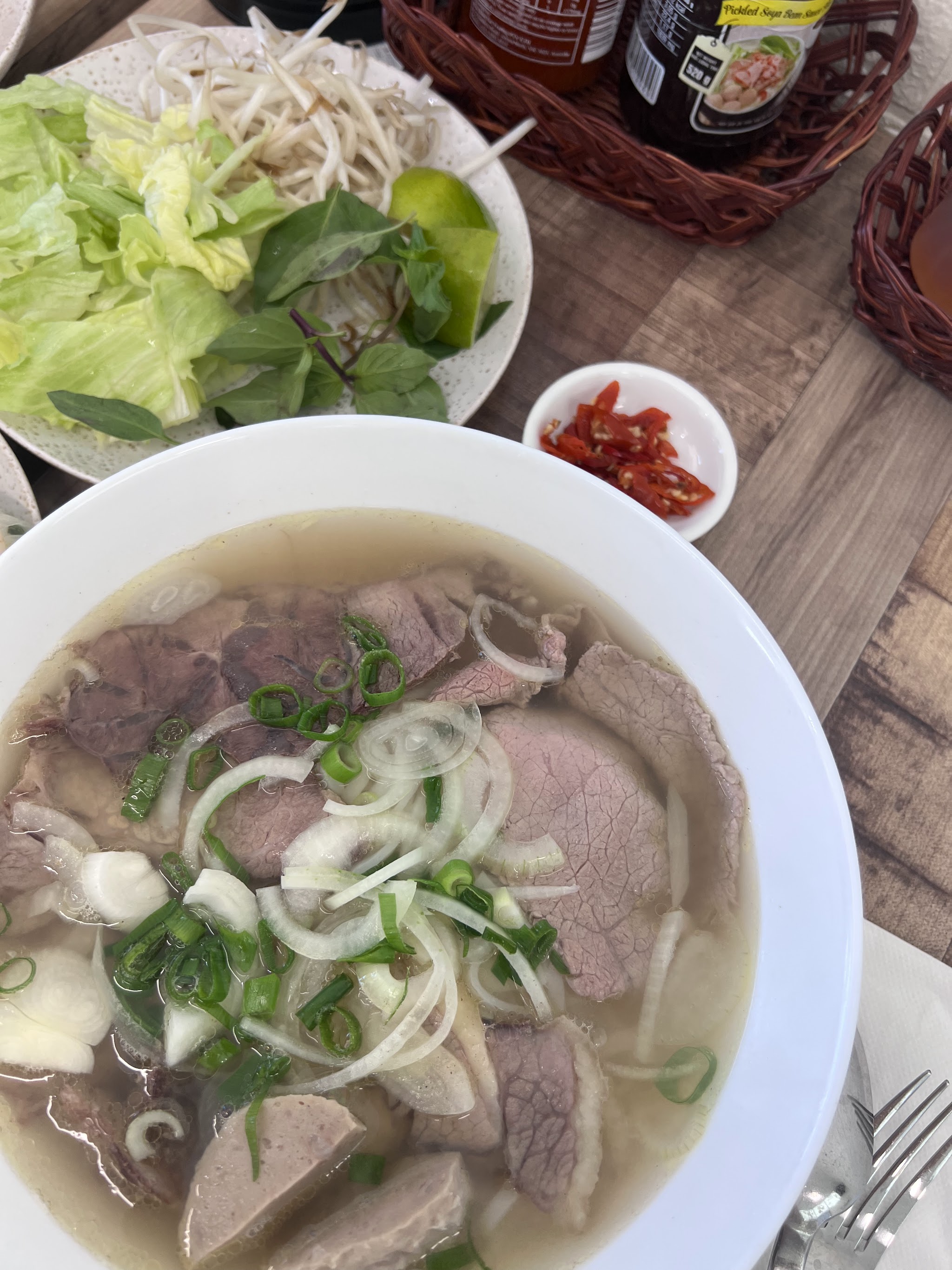 A bowl of beef phở with a side plate of fresh herbs, lime and chilli